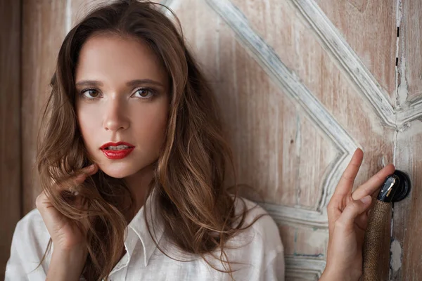 Retrato cercano de una hermosa joven con labios rojos en una camisa blanca. una mano de la chica está en la cara, la otra mano está sosteniendo el mango de la puerta de madera —  Fotos de Stock