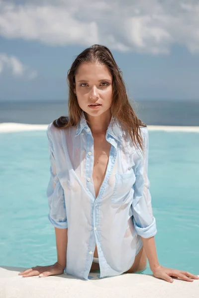 Una hermosa joven modelo bronceada con una camisa azul mojada apoya sus manos en el borde de la piscina. piscina infinita azul y vista al mar en el fondo — Foto de Stock