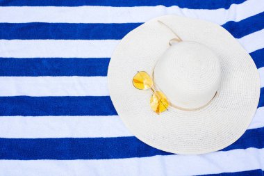 Beyaz bir şapka, sarı güneş gözlüğü ve bir bardak su mavi ve beyaz çizgili havlu üzerinde yatıyor. yukarıdan görünüm