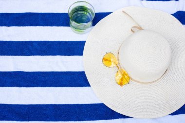 Beyaz bir şapka, sarı güneş gözlüğü ve bir bardak su mavi ve beyaz çizgili havlu üzerinde yatıyor. yukarıdan görünüm