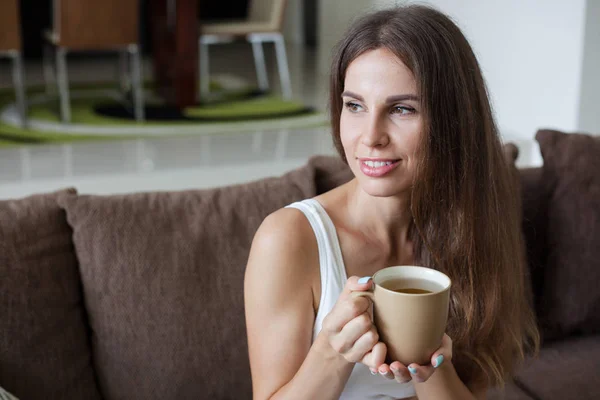 Porträtt av vackra leende brunett flicka sitter på soffan i vardagsrummet och innehar mugg te i hennes händer. Flickan tittar bort — Stockfoto