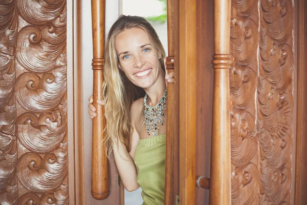 Hermosa chica rubia sonriente con el pelo largo asoma a través de una puerta de madera abierta — Foto de Stock