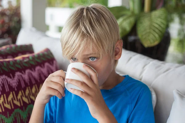 Mavi tişörtlü yakışıklı sarışın bir genç kanepede oturur ve beyaz bir bardaktan içer. çocuk mesafeye bakar