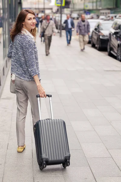 vertical portrait of beautiful smiling elegant young woman in business clothes walks along the street along a glass building and rolls a small suitcase on wheels