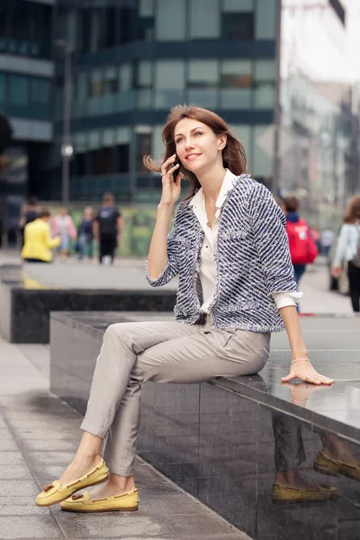 attractive smiling young business brunette girl dressed in elegant clothes sits on a bench on a street in the city center and talking on mobile phone. Glass building in the background