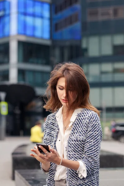 beautiful young business brunette woman dressed in elegant clothes sits on a bench on a street in the city center and talking on a cell phone. Glass building in the background