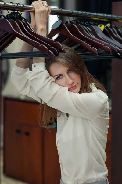 nothing to wear. Beautiful serios young brunette woman in elegant light shirt and pants stands in the dressing room and holds on to floor hanger racks