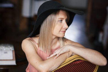 Kül gül renkli elbise ve siyah keçe şapka çiller ile güzel bir dalgın genç sarışın kadının yakın sıcak romantik portre. Kız içeride oturuyor ve yan tarafa bakıyor..