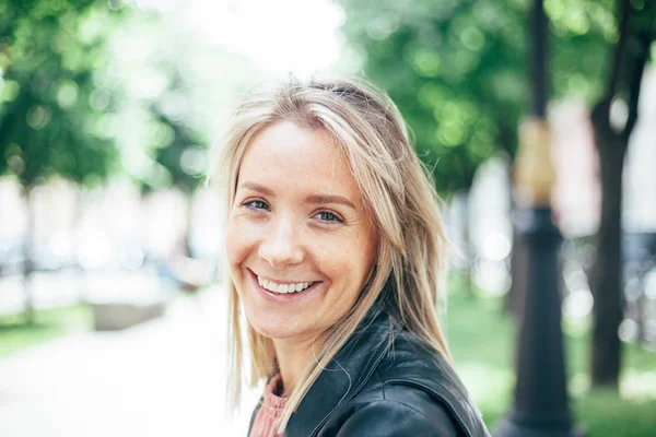 Retrato de cerca de una hermosa rubia sonriente con pecas. Al fondo una avenida con árboles en desenfoque —  Fotos de Stock