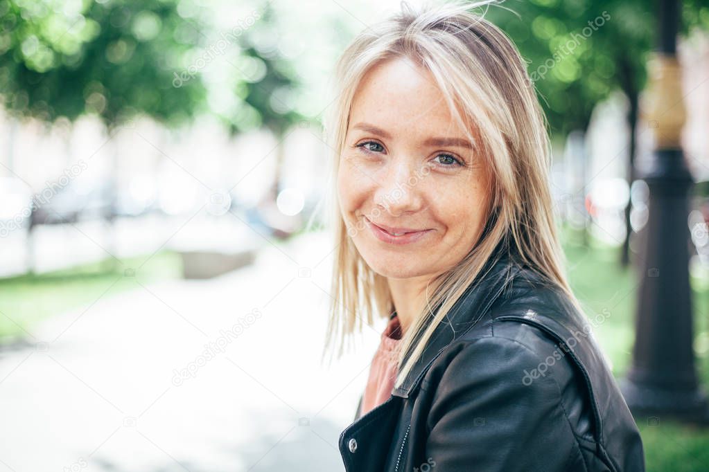 Closeup portrait of a smiling happy beautiful young woman blonde with sunspots. In the background an avenue with trees in defocus