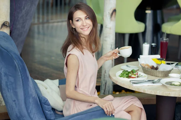Hermosa joven feliz sonriente en un vestido rosa tiene almuerzo en un restaurante. Comida y bebidas en la mesa . —  Fotos de Stock