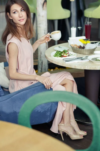 Hermosa mujer joven en un vestido rosa tiene almuerzo en un restaurante. Un plato con ensalada en la mesa. Chica está sosteniendo la taza de capuchino y mirando hacia otro lado —  Fotos de Stock