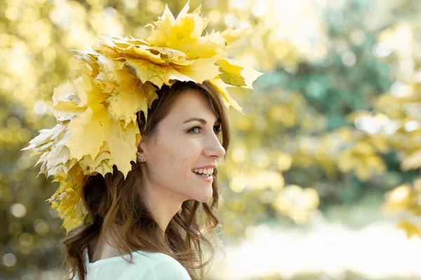 Retrato de cerca de una hermosa morena feliz con una corona de hojas de arce amarillo en la cabeza. Follaje dorado otoñal de árboles en el fondo —  Fotos de Stock