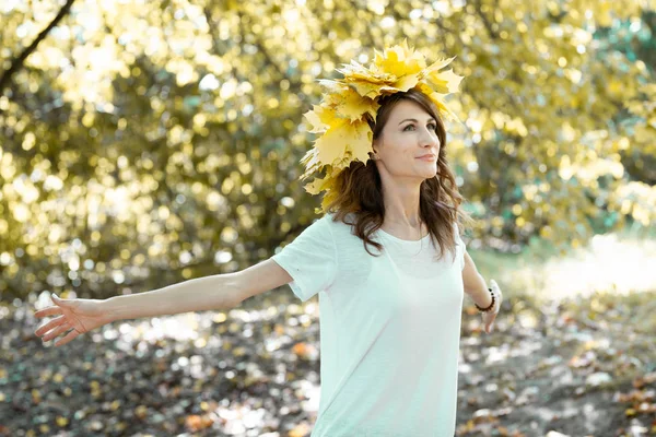 Retrato de cerca de una atractiva joven morena sonriente con una corona de hojas de arce amarillo en la cabeza. chica con los brazos extendidos a los lados. Bosque dorado de otoño en el fondo —  Fotos de Stock