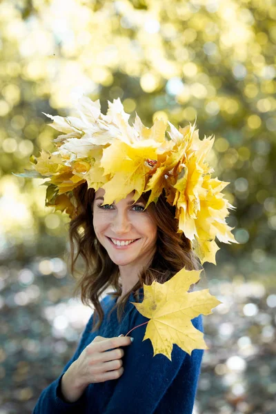 Retrato de una hermosa chica morena sonriente con una corona de hojas de arce amarillo en la cabeza y en jersey azul. niña tiene hoja de arce de otoño en la mano. Parque de oro en el fondo . — Foto de Stock