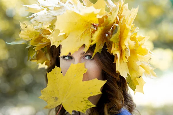 Retrato de cerca de una hermosa joven morena con una corona de hojas de arce amarillo en la cabeza. chica cubre toda su cara con una hoja de arce amarillo, excepto los ojos. Parque de oro en el fondo . — Foto de Stock