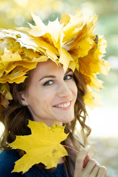 Retrato de cerca de una hermosa joven morena sonriente y feliz con guirnalda de hojas de arce amarillo en la cabeza y en jersey azul. niña tiene hojas de arce de otoño en la mano. Parque de oro en el fondo . — Foto de Stock