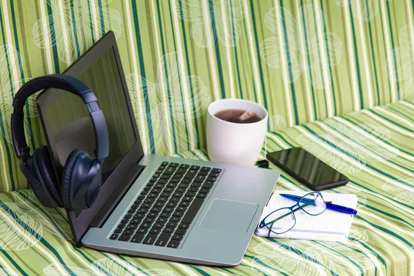 Büyük kulaklıklı dizüstü bilgisayar, cep telefonu, çay fincanı, dışarıdaki yeşil çizgili şilte üzerinde kalem olan defter.
