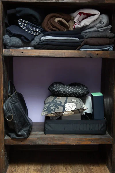 Деревянные полки с кучей сложенной одежды разных темных цветов, черный старый кожаный портфель, подушки, коробки и другие вещи — стоковое фото