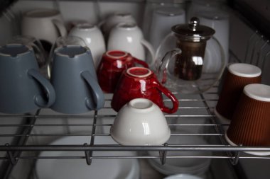 Bulaşık kurutma makinesinin metal ızgaralarında bir sürü seramik, cam bardak, bardak ve farklı renkte bardaklar var.