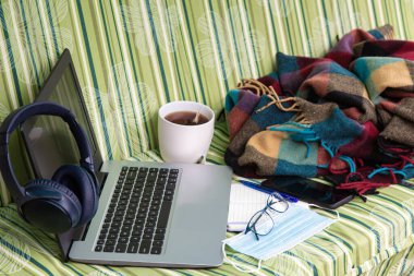 Bilgisayar, ekose, çay fincanı, kulaklık, tıbbi maske, parlak yeşil koltukta gözlük. uzak çalışma, tecrit hayatı