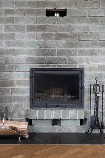 gray brick wall fireplace with traces of black soot. cast-iron fireplace accessories and firewood in a metal basket nearby.