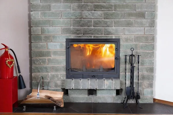 front view of a gray brick fireplace with a blazing fire behind a glass door. a red fire extinguisher, firewood and cast-iron accessories near the hearth. Fire safety. copy space