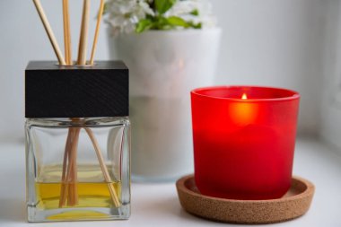 Siyah kapaklı, ahşap sopalı ve sarı aromatik yağlı yakın plan cam difüzörü. Kırmızı camlı mum. Beyaz duvar ve arka planda beyaz çiçekler olan beyaz cam. seçici odak