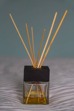Siyah ahşap kaplamalı bir cam dağıtıcı, tahta sopalar ve gri bir yatak örtüsünün üzerinde sarı aromalı yağ standı. Arka planda gri-mavi duvar var. seçici odak