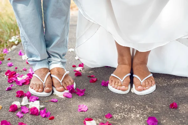 close-up of feet of bride and groom in white flip flops outside. Pink rose petals on the ground. The woman lifts her white wedding dress. No face