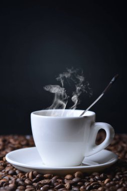 Bir fincan sıcak kahve masanın üzerinde dağınık kahve çekirdeklerinin üzerinde, fincandan buhar çıkıyor, koyu arka plan.