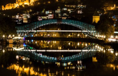 Tiflis 'in merkezindeki dostluk köprüsünde gece görüşü. Alacakaranlık, suda ışık yansımaları ve antik şatoda ve arka planda küçük evler.