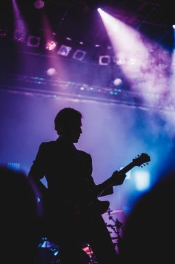 Sahnede gitar çalan bir adamın silueti. Koyu arkaplan, duman, spot ışıkları