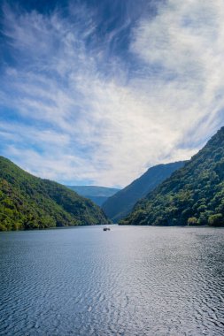 Lugo, İspanya 'daki Ribeira Sacra' da Sil nehrinin kanyonu. Arka planda bir tekne ve bulutlu bir gökyüzü olan teraslı üzüm bağlarıyla dağlar arasında nehir manzarası.