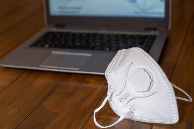 KN95 FPP2 Corona Virüs Koruma Maskesi ahşap bir masanın üzerinde ve arka planda odakları bozuk bir laptop var.