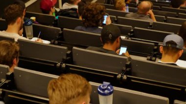 Öğrenciler konferans salonunun önünde öğretmeni dinliyorlar.