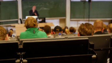 Öğrenciler konferans salonunun önünde öğretmeni dinliyorlar.