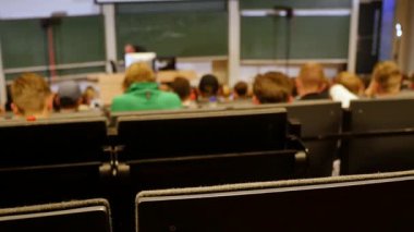 Öğrenciler konferans salonunun önünde öğretmeni dinliyorlar.