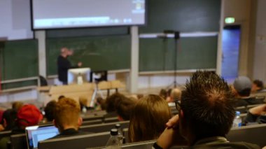 Öğrenciler konferans salonunun önünde öğretmeni dinliyorlar.