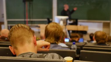 Öğrenciler konferans salonunun önünde öğretmeni dinliyorlar.