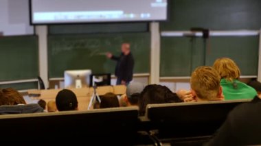 Öğrenciler konferans salonunun önünde öğretmeni dinliyorlar.