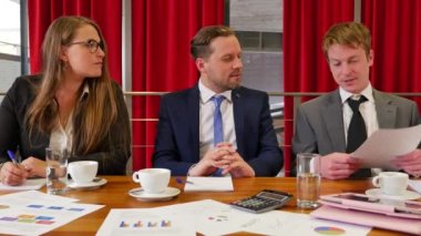 Üç iş ortağı konferans salonunda buluşuyor ve arka planda kırmızı perdeler var.