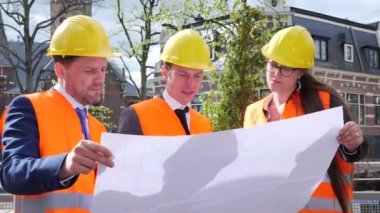 Miğferli üç mimardan oluşan ekip proje planını tartışıyor.