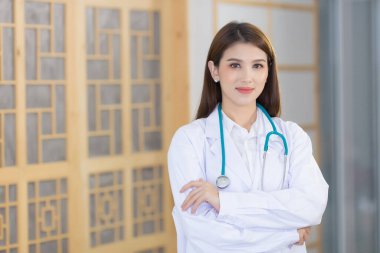 Genç ve güzel Asyalı kadın doktor kollarını kavuşturup mutlu mesut ayakta duruyor ve hastanede gülümsüyor. Beyaz bir bornoz ve steteskop giyiyordu..