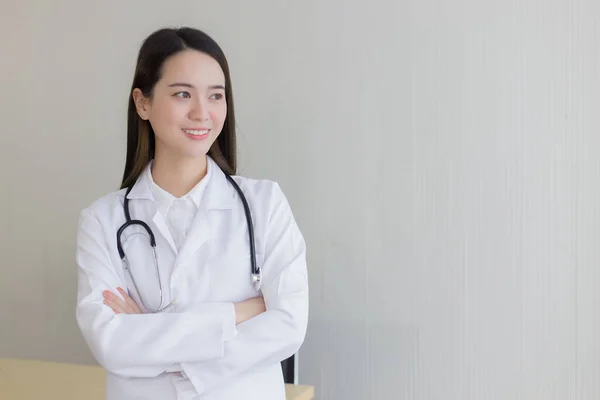 Asian female doctor with black long hair wears a white lab coat and stethoscope. She stands and cross arms in office at hospital.