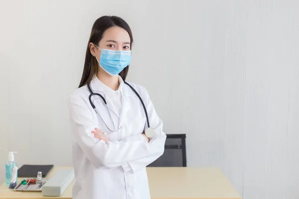 黒い長い髪をしているアジア系女性の医師は 医療用フェイスマスクを着用し 病院のオフィスで腕を組んでいる間 一様として聴診器と白衣を着用する 病院で医療面マスクを着用する — ストック写真