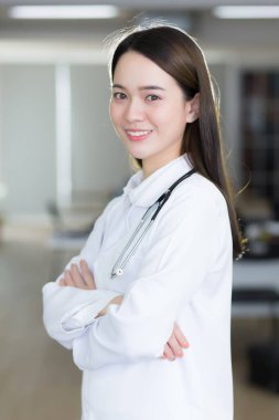 Asyalı kadın doktor kendine güvenerek ayakta duruyor ve gülümsüyor. Hastanede beyaz laboratuvar önlüğü ve steteskop giyerken kol kola giriyor..