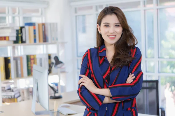 An Asian confident businesswoman is standing with arm crossed and smiling in a working room at home (work from home concept).