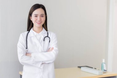 Asyalı kadın doktor kollarını kavuşturup mutlu mesut ayakta duruyor ve hastanede gülümsüyor. Beyaz bir bornoz ve steteskop giyiyordu..