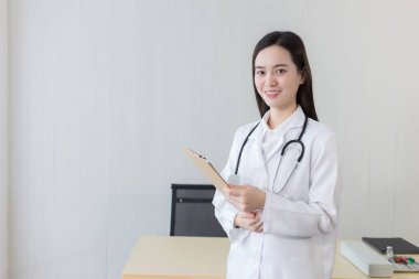 Hastanede çalışan genç, güzel Asyalı kadın doktor. Beyaz bir bornoz ve steteskop takıyor ve elinde bir pano tutuyor. Sağlık ve Corona virüs kavramı.